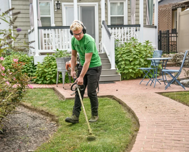 Crew member in a green shirt trimming a lawn edge along a brick path